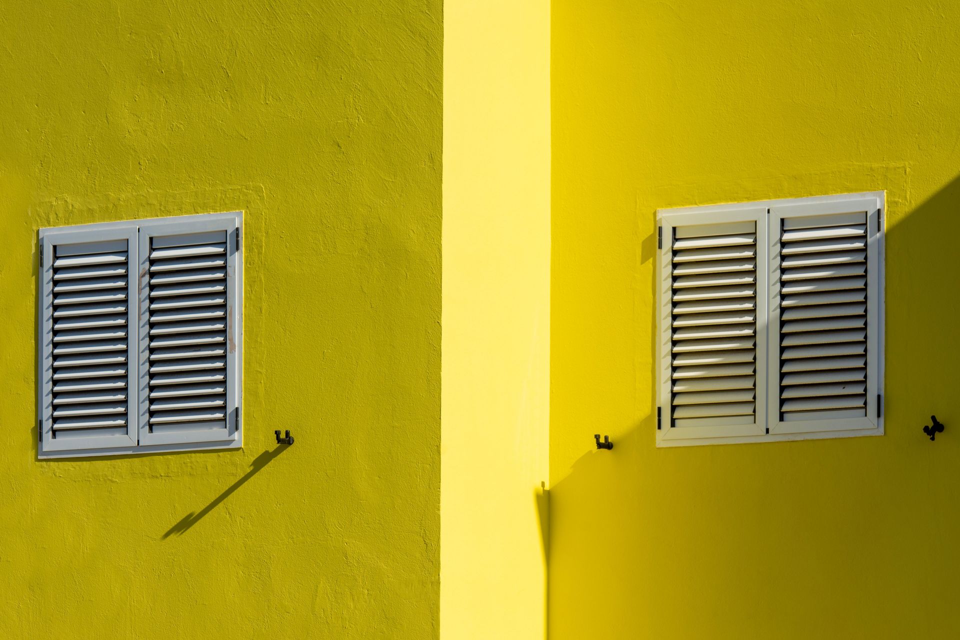 Close-up of a vibrant yellow building facade with white window shutters casting shadows.