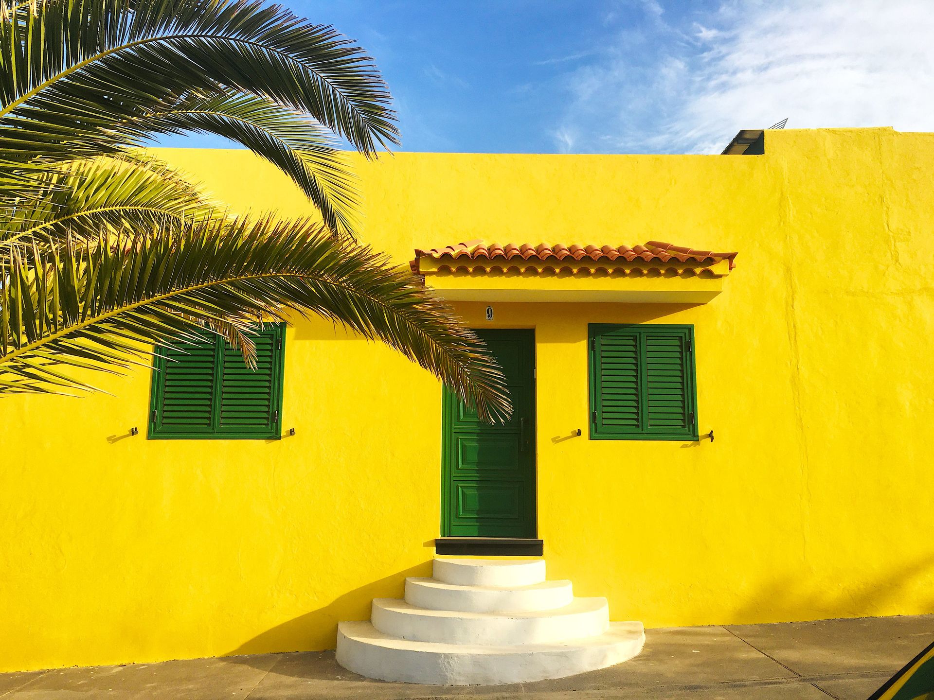 Vibrant yellow house facade in Spain with green shutters and a palm tree.